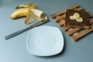 A delicious piece of toast with chocolate spread and banana slices on a modern kitchen countertop with a kitchen knife, plate and vintage wooden surface
