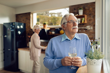 Senior man with wineglass day dreaming while his wife is preparing food in the kitchen.