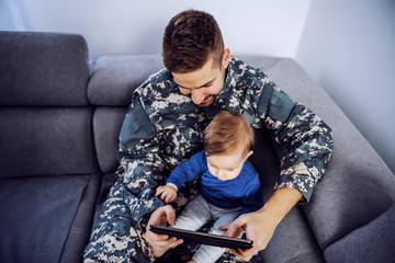 Top view of brave soldier making up for the lost time with his son. Man holding toddler in lap, tablet in hands and watching cartoons.