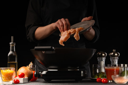 The chef pours the chicken fillet into a wok. Freezing in motion. Restaurant menu, cooking. On a black background, photo for advertising or design, restaurant business, cooking and recipe book