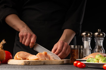 Chef slices chicken, for cooking, and salad. Against the background of vegetables, cooking and home recipe book