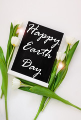 Happy Earth Day April 22 greeting message sign on black chalk board with tulip flowers on white background. World ecology concept card.Saving environment, save clean planet.Blackboard with text,banner