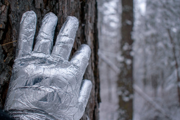 Hand in a shiny metal glove rests on a tree. Forest in blur, winter, snow. The concept of an extraterrestrial creature, an alien, an ice king, a knight.