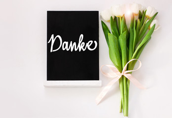 Danke thank you in German card sign on black chalkboard with tulip flowers on white background flat lay. Blackboard greeting text message top view. Natural floral green leaves. Thankful quote banner.