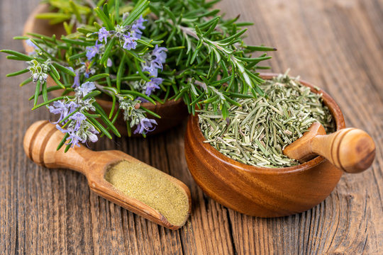 A wooden bowl with blooming and fresh rosemary twigs, a wooden bowl with whole dried rosmary and a spice shovel with ground rosemary on a rustic wooden background