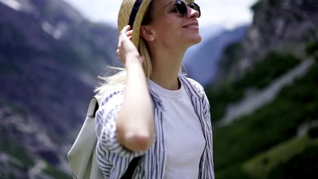 Happy female traveler in hat and sunglasses raising hands while feeling freedom of nature in mountains, excited hipster girl wanderlust satisfied with summer journey to alps. Celebrating success 
