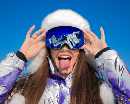 Funny portrait of a young woman in a ski goggles showing tongue on a ski resort.