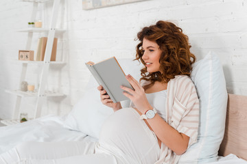 Attractive pregnant woman smiling while reading book on bed