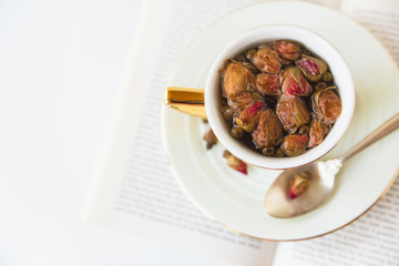 Herbal tea dried roses in cup and spoon.Hot healthy drink on the open book.White background.Minimalist style.Copy space.Top view.