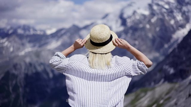 Back view of young woman standing on wind with raising hands up enjoying nature and freedom atmosphere. Carefree hipster girl wanderlust looking at snowy mountains inspiring at vacations summer travel