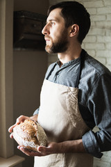 Bearded young man in rustic apron holding a loaf of bread in his hands