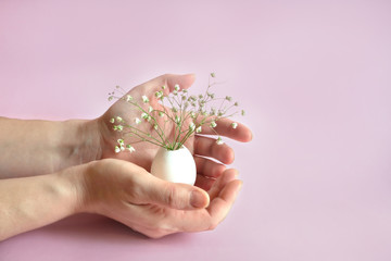 Hands holding a white egg with a bouquet of flowers from the gypsophila. The concept of the celebration of Easter. Delicate solid pink background. Minimalism, copy space.Tender postcard.
