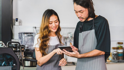 Young Asian man and woman barista wear apron using tablet received order online at bar counter in coffee shop with smile face.Concept of cafe and coffee shop small business.Vintage tone