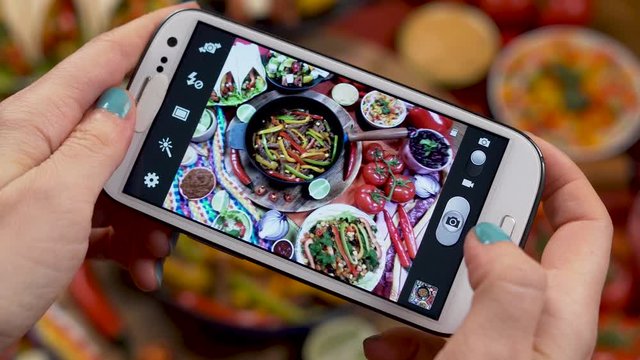 A Woman's Hands Take Photos Of Traditional Mexican Food with a Smartphone Hot Fajitas is Served with Sauces and Tortilla and Vegetables, Burritos, Bean Soup with Chili.