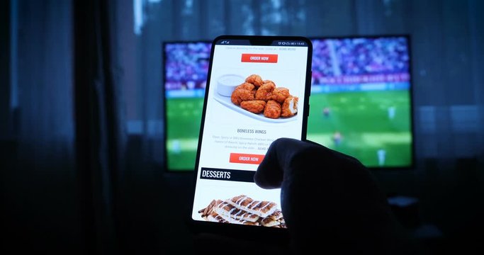 Background of the TV at home (at the bar), the guy orders different snacks, pizza and sweets scrolling through internet stores using mobile phones. Concept: Sport.