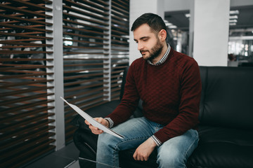 A brunette bearded man sits on a couch and reads a contract in a saloon. Happy, worried, skeptic,...