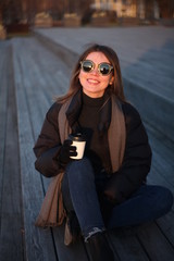 Young beautiful girl in sunglasses and a cap sits on the street and drinks coffee