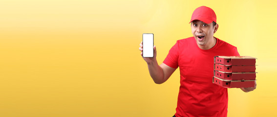  handsome Asian man in red cap, giving food order italian pizza in cardboard  boxes isolated on yellow background in studio With copy space. holding mobile phone with blank  white  empty screen. 