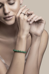 Cropped portrait of a charming lady with golden earrings, silver rings and a gemstone bracelet made of green beads. The semi-naked girl with crossed hands is posing on the beige background.  - obrazy, fototapety, plakaty