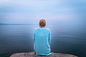 Fototapeta premium Beautiful single solo young traveler blond woman with tranquit jacket sitting alone on stone looking in to water in the blue ocean foggy mist lake in cold winter with sad dark depression emotional
