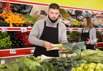 Male shop assistant lays fresh leek on counter in grocery shop