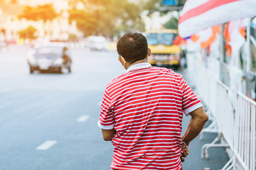 Back view of male patient with mask in red and white shirt standing at bus stop and wait for taxi or bus in the city to go to the hospital.