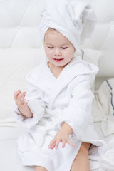 Little cute girl sitting on the bed in a white bathrobe and a towel on her head painting with varnish nails on her hands. Little girl doing herself a manicure
