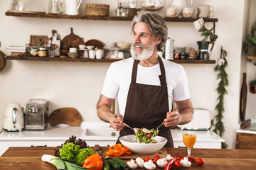 Image of happy gray-haired man in apron smiling and preparing salad
