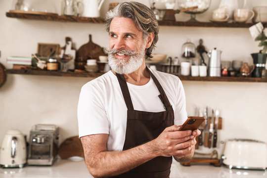 Image of cheerful mature man in apron smiling and using cellphone