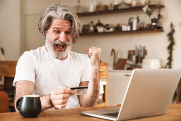Image of excited mature man working with laptop and holding credit card