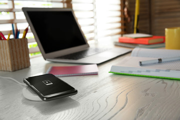 Mobile phone with wireless charger on white wooden table. Modern workplace accessory