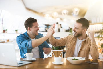 two colleagues men giving each other high five looking winning working on successful common project during lunch break at cafe, multitasking and teamwork concept