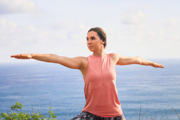 Close up warrior pose. Young woman practicing yoga, standing in Virabhadrasana pose. Outdoor yoga on the cliff. Bali, Indonesia