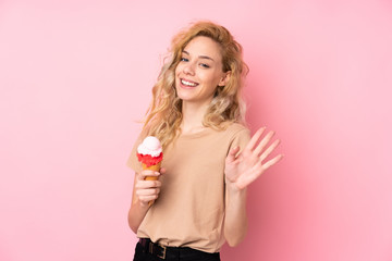 Young blonde woman holding a cornet ice cream isolated on pink background saluting with hand with happy expression