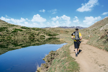 A man makes trekking walking along a road with a lake next to it and mountains in the background on a sunny day