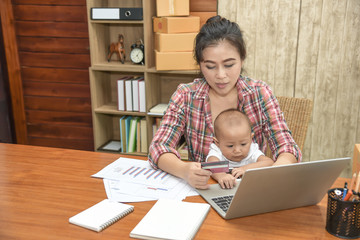 Pretty young single mom working at home on a laptop computer while holding her baby girl sitting on her lap enjoying watching mother working.
