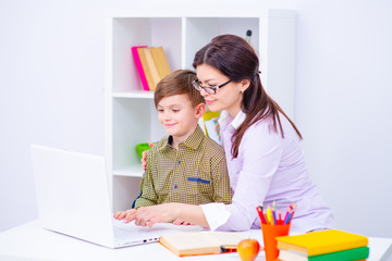 A boy sitting at a table at home in front of a laptop. A teacher sitting next to a boy helping him to work with a computer