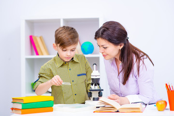 A boy sitting at a table at home in front of a microscope. A teacher sitting next to a boy helping...