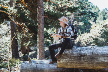 A red-haired woman with a hat and backpack, sightseeing through a forest looking at a map sitting on a tree trunk
