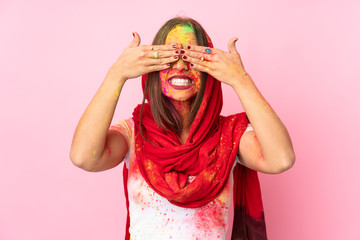Young Indian woman with colorful holi powders on her face isolated on pink background covering eyes by hands and smiling