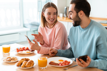 Obraz na płótnie Canvas Portrait of young joyful couple using cellphones while having breakfast