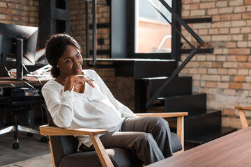 smiling, pregnant african american businesswoman sitting in armchair and looking away