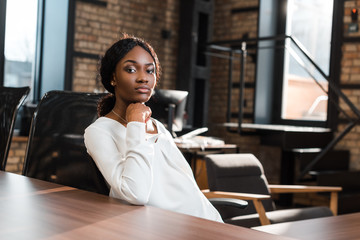 thoughtful, pregnant african american businesswoman sitting at office desk and looking at camera
