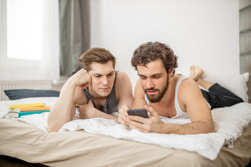 young caucasian gays lie on bed with smartphone in hands. homosexual happy male couple, young...