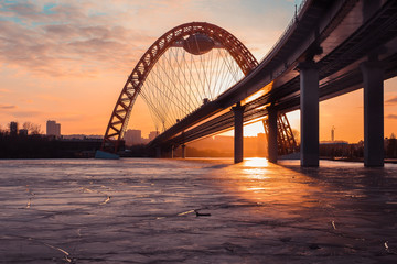 The photo shows picturesque bridge with big red arch over the river. This cable-stayed bridge stands on the frozen Moscow river. Crimson sunny rays illuminate bridge details and ice on the sunset. 