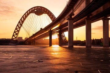 Obraz premium The photo shows picturesque bridge with big red arch over the river. This cable-stayed bridge stands on the frozen Moscow river. Crimson sunny rays illuminate bridge details and ice on the sunset. 