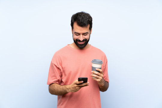 Young man with beard holding a take away coffee over isolated blue background sending a message with the mobile