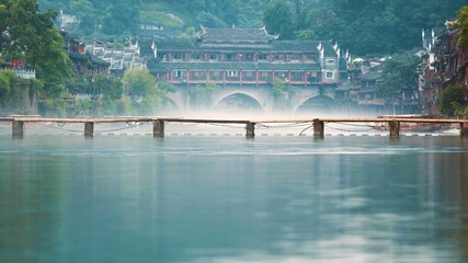 Fototapeta na wymiar A simple wooden bridge on a river canal with buildings in the background. Fenghuang County, Hunan, China