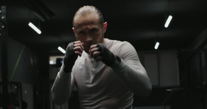 Martial arts sportsman in grey training suit, boxing at camera in close-up, against dark gym interior