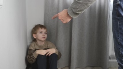 A frightened, crying child is sitting in the corner of the room. Violence against children in the family. The male father holds the belt in his hand.